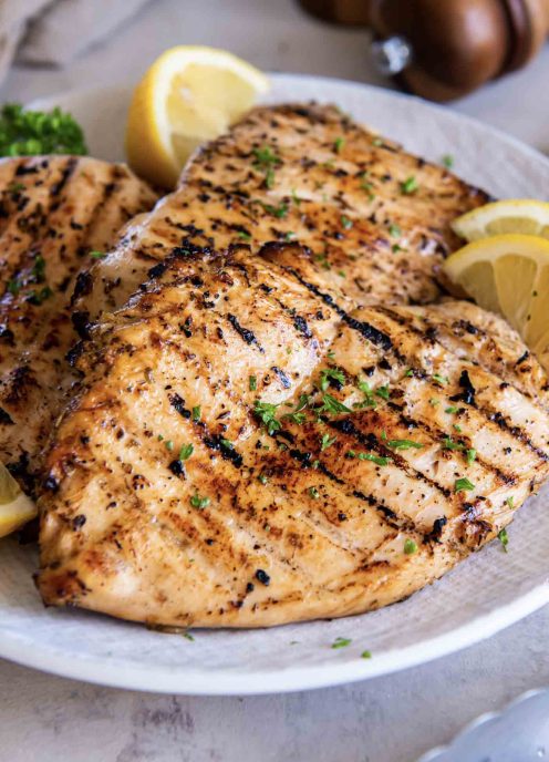 Three grilled chicken breasts on a plate garnished with lemon wedges.